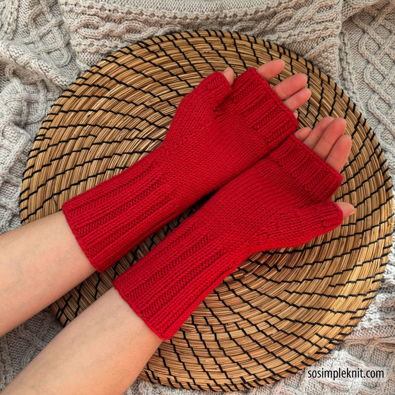 The image showcases a pair of vibrant red fingerless gloves laid on a woven straw mat. The gloves feature a ribbed texture and extend to the mid-forearm. In the background, a light gray knitted fabric adds a cozy, soft contrast, enhancing the focus on the gloves. The hands completing the display are lightly tanned, positioned with palms facing upwards, conveying a relaxed and inviting presentation.