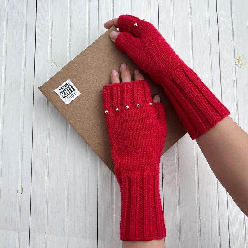 The image shows a pair of bright red fingerless gloves displayed on a kraft cardboard package. The gloves have ribbed cuffs and are adorned with small silver beads along the wrist. A hand is positioned beneath the gloves, while another hand lightly touches the cardboard. The background features white wooden planks, creating a clean and simple aesthetic.