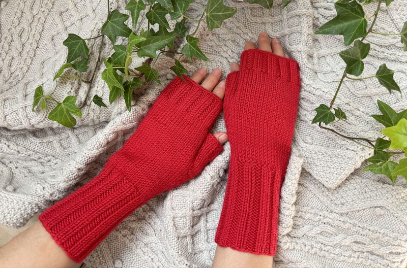 A pair of red fingerless gloves lies on a light gray knitted background, surrounded by green ivy leaves. The gloves are ribbed, with the fingers cut off, showcasing a textured design. The lighting is soft, highlighting the stitches and contrasting the red color against the neutral background.