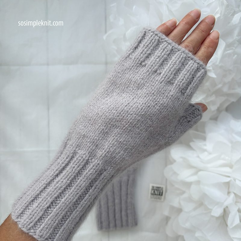 A close-up image of a hand wearing a light gray fingerless glove with ribbed detailing. The glove is knitted and soft-looking, showcasing a smooth texture. In the background, a white, soft fabric is draped under the hand, with subtle floral decorations. The overall composition centers on the glove and hand, emphasizing the knitting pattern and style.