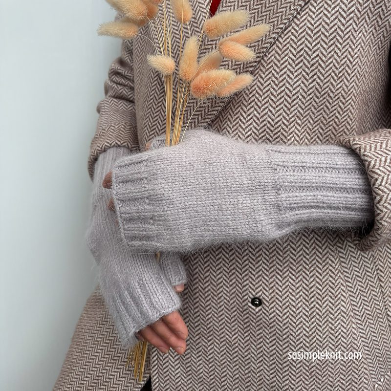 The image features a close-up of a person wearing light gray fingerless gloves, structured with ribbed detailing. The gloves have cut-outs for the fingers, showcasing a relaxed and cozy fit. The person's arms are framed against a neutral background, and they hold a bouquet of dried flowers. The individual is dressed in a patterned brown coat with a herringbone design, which adds texture to the composition.