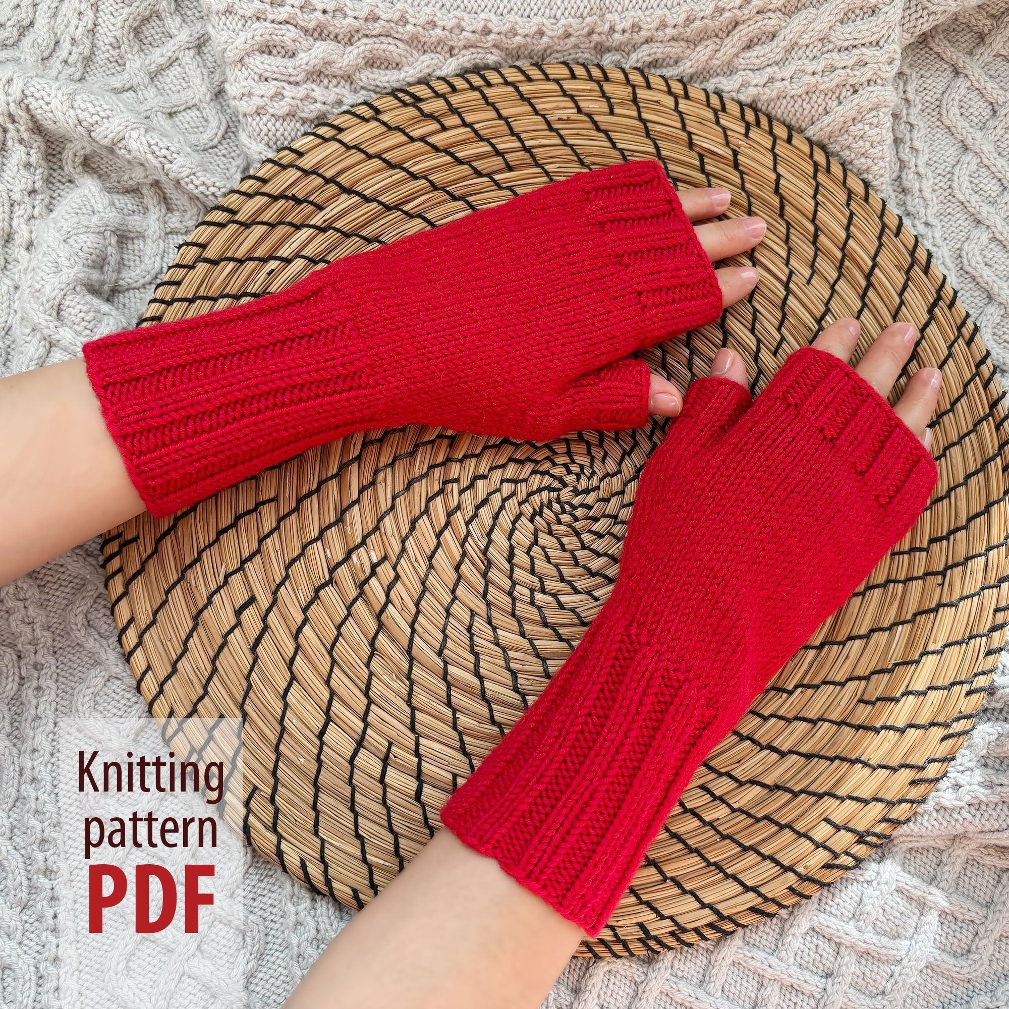 Detailed image of two hands in bright red knitted fingerless gloves. The hands rest on a textured woven tray on top of a knitted blanket. A text overlay is visible: "Knitting pattern PDF"