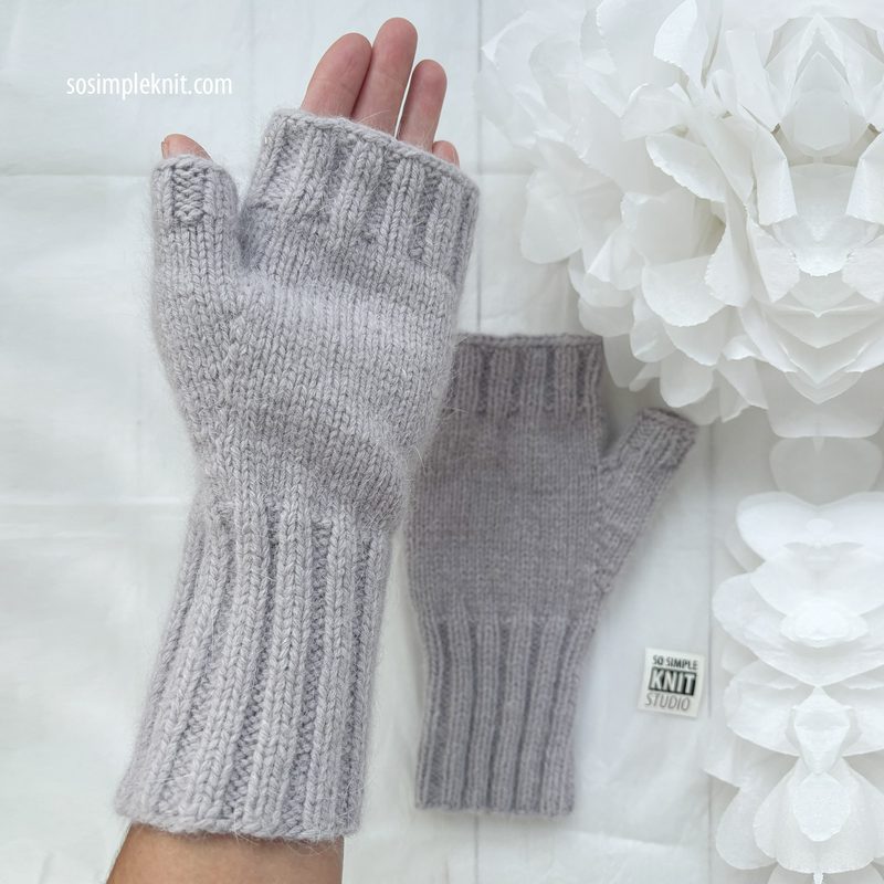 The image shows a pair of light gray fingerless gloves displayed on a neutral background. The gloves are ribbed at the wrist and feature a smooth knit texture, with fingers missing to allow for free movement. One glove is laid flat, while the other is held up by a hand. Behind the gloves, there is a white decorative element that resembles a cluster of paper flowers, creating a soft and delicate contrast. The overall composition focuses on the gloves, highlighting their design and color.