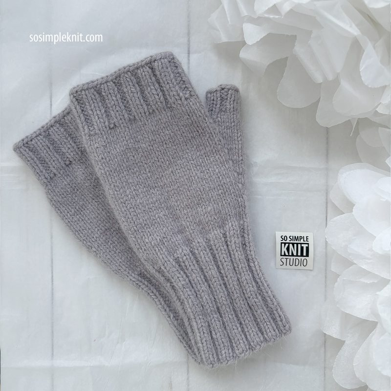 The image features a pair of light gray fingerless gloves knitted in a ribbed pattern, displayed on a textured white surface. The gloves have a snug fit with a ribbed cuff at the wrist. In the background, soft white paper flowers are partially visible, adding a delicate touch to the composition. A small logo tag reading "SO SIMPLE KNIT STUDIO" is placed between the gloves and the bottom edge of the image.