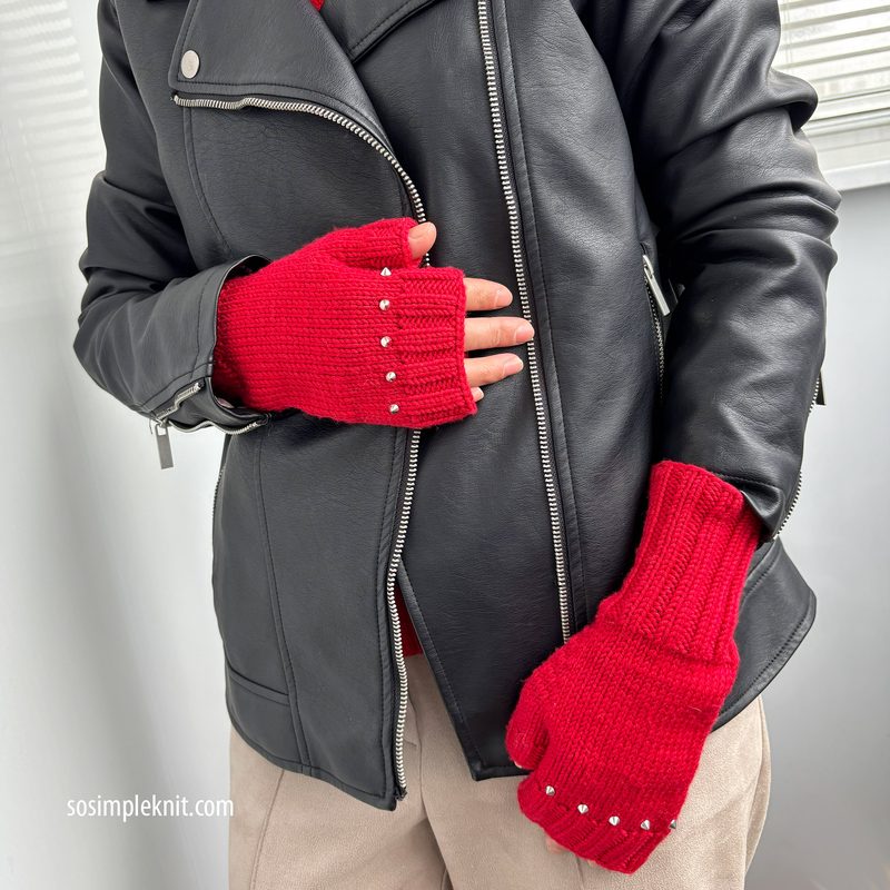 The image features a pair of red fingerless gloves worn by a person whose hands are positioned near their body. The gloves have a ribbed texture and are adorned with small silver studs along the edges. The individual is wearing a black leather jacket, partially unzipped, and beige pants. The background appears light-colored, and the overall lighting is bright, highlighting the gloves and jacket.