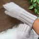 A close-up image of a pair of light gray fingerless gloves made from a soft, knitted fabric. The gloves feature ribbed cuffs and a smooth body, partially covering a hand and wrist. The background consists of textured brown fabric and green foliage, creating a natural and cozy atmosphere. The gloves are positioned at an angle to show their shape and texture effectively.