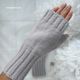 A close-up image of a hand wearing a light gray fingerless glove with ribbed detailing. The glove is knitted and soft-looking, showcasing a smooth texture. In the background, a white, soft fabric is draped under the hand, with subtle floral decorations. The overall composition centers on the glove and hand, emphasizing the knitting pattern and style.