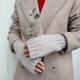 A pair of light gray fingerless gloves are worn by a person with their hands clasped together, partly concealed by the sleeves of a beige herringbone coat. The gloves have a ribbed texture and fit snugly at the wrists. The person is wearing a deep red top underneath the coat, and a small branch of eucalyptus is held between their fingers. The background is plain and minimal, highlighting the gloves and the person’s outfit.