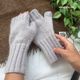 The image shows a pair of light gray fingerless gloves made from a soft, textured yarn, displayed against a brown woven background. A hand is positioned below the gloves, partially holding them, while a small label with "SO SIMPLE" is visible near the wrist area. Green leaves from a nearby plant are also included in the frame, contrasting with the neutral colors of the gloves and background.