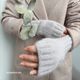 A close-up image showing a pair of light gray fingerless gloves being held by a hand with pale skin, featuring a ribbed texture and an open design at the fingertips. The gloves are worn by a person dressed in a soft, neutral-toned, patterned coat. The other hand holds a single sprig of eucalyptus, adding a touch of greenery to the composition. The background is softly blurred, focusing on the gloves and the hand.