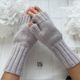A close-up image of a pair of light gray fingerless gloves made from a soft, textured yarn. The gloves feature a ribbed design and are worn on two hands, positioned one above the other against a light-colored surface. In the background, there are two white faux flowers, creating a soft and neutral aesthetic. The branding label "SO SIMPLE KNIT STUDIO" is visible on the surface below the gloves.