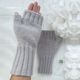 The image shows a pair of light gray fingerless gloves displayed on a neutral background. The gloves are ribbed at the wrist and feature a smooth knit texture, with fingers missing to allow for free movement. One glove is laid flat, while the other is held up by a hand. Behind the gloves, there is a white decorative element that resembles a cluster of paper flowers, creating a soft and delicate contrast. The overall composition focuses on the gloves, highlighting their design and color.