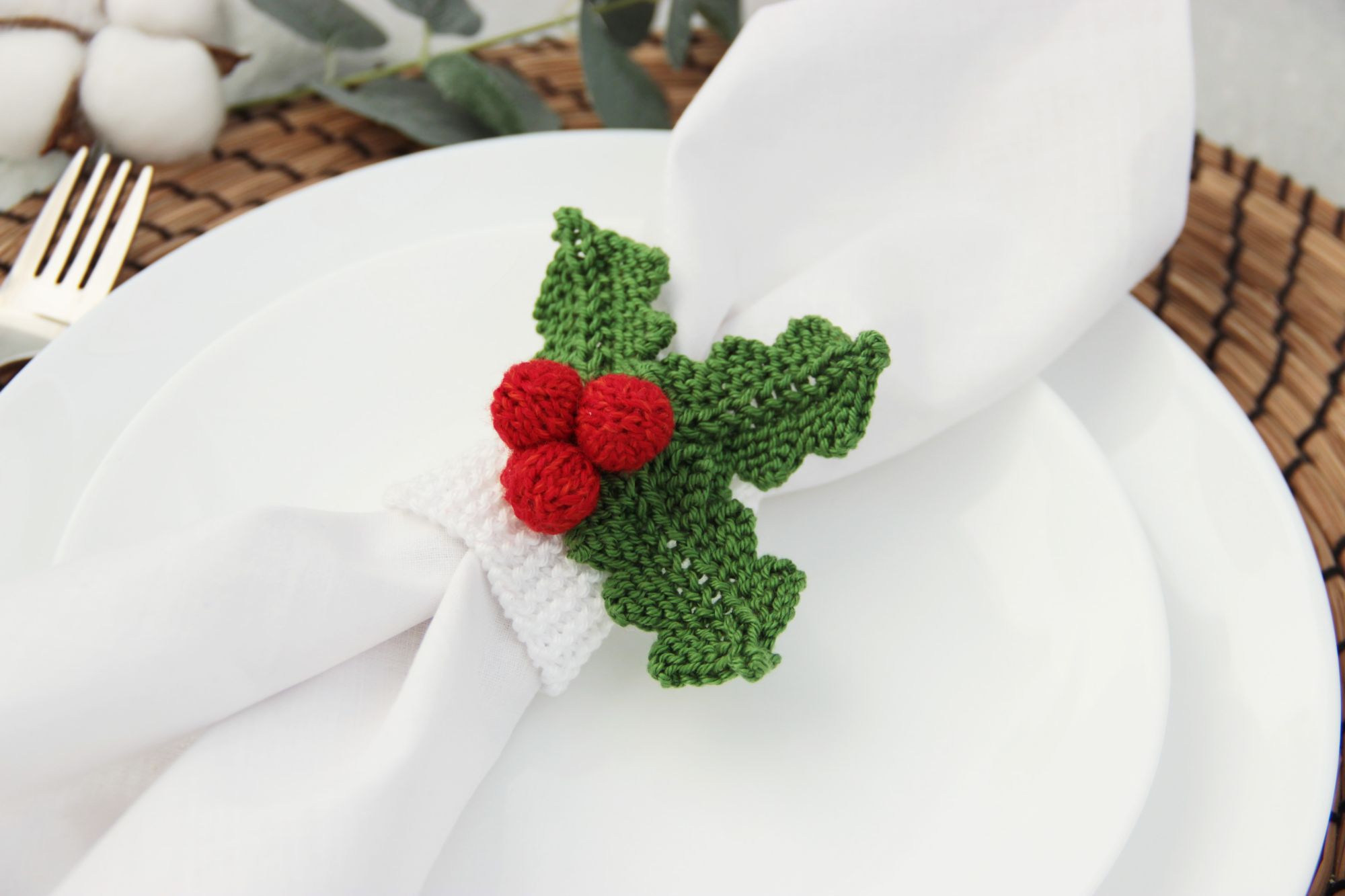 A knitted napkin ring in the shape of a Christmas holly leaf, with green leaves and red berries