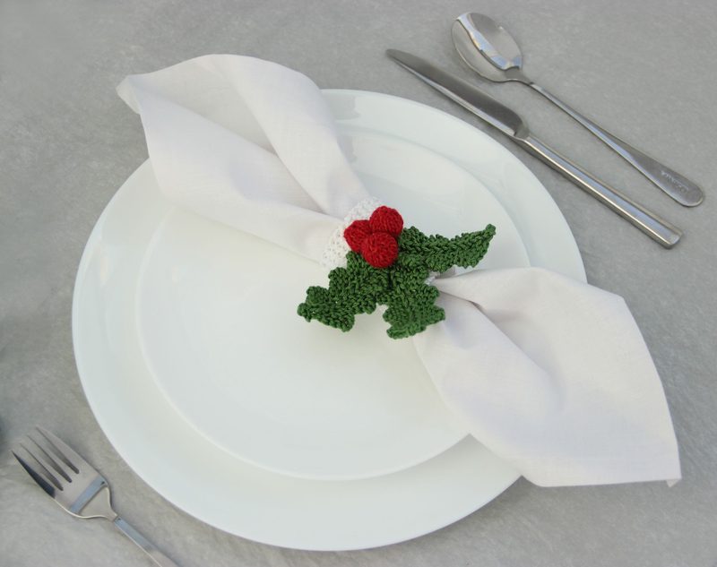 A white dinner plate set on a gray surface features a folded white napkin held in place by a knitted holly napkin ring. The napkin ring is decorated with red berries and green leaves, showcasing intricate knitting patterns. A fork and knife are positioned alongside the plate, completing the dining setting. The overall style is festive and elegant, suitable for a holiday table setting.