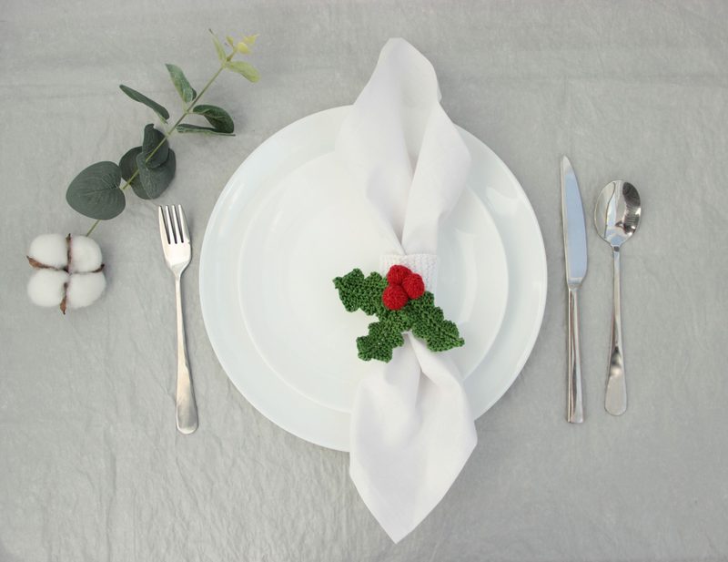 A white plate is set on a textured, gray surface, with a neatly folded white napkin secured by a green knitted holly napkin ring featuring two red berries. To the left, a silver fork is placed beside the plate, and to the right, a knife and spoon are arranged. A small cotton boll with fibers is positioned near the bottom left, and a sprig of green leaves adds a natural touch beside the plate.