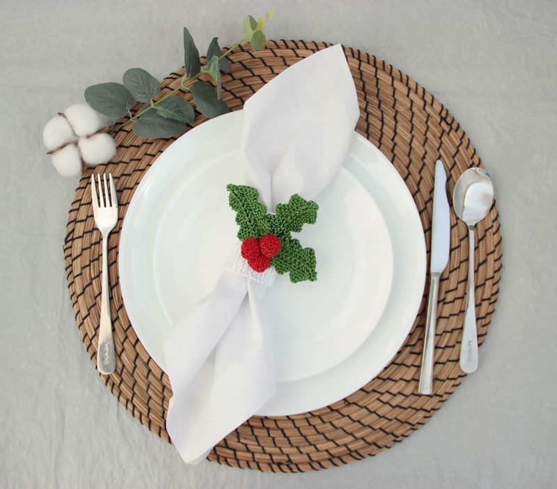 A white dinner napkin is elegantly folded and secured with a knitted holly napkin ring featuring green leaves and a red berry. The napkin rests on a white plate, which is set on a woven straw placemat. To the left, a sprig of eucalyptus and a small cluster of cotton bolls add a natural touch, while a fork and knife are positioned beside the plate, creating a festive table setting. The overall ambiance is warm and inviting, with subtle, earthy tones and textures.