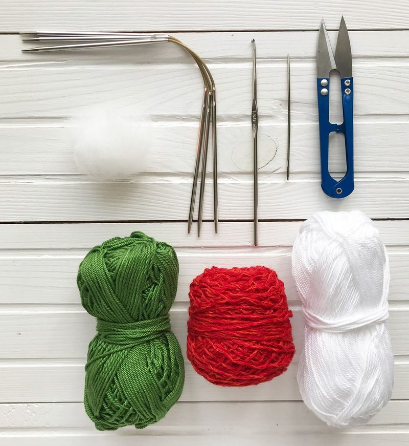 The image features a flat lay arrangement on a white wooden surface. It includes three skeins of yarn in green, red, and white colors, positioned horizontally. Above the yarn, there are knitting needles, a crochet hook, a sewing needle, a pair of scissors, and a stitch gauge tool, all arranged in a tidy manner. The yarn is soft and textured, while the tools are metallic and colorful.