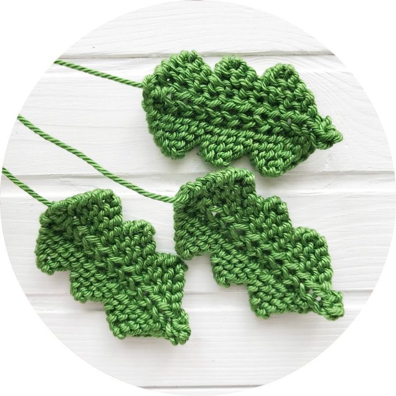 A circular image featuring three knitted leaf-shaped napkin rings in various shades of green, creatively arranged with thin green yarn connecting them. The background is a light textured surface, enhancing the focus on the napkin rings.