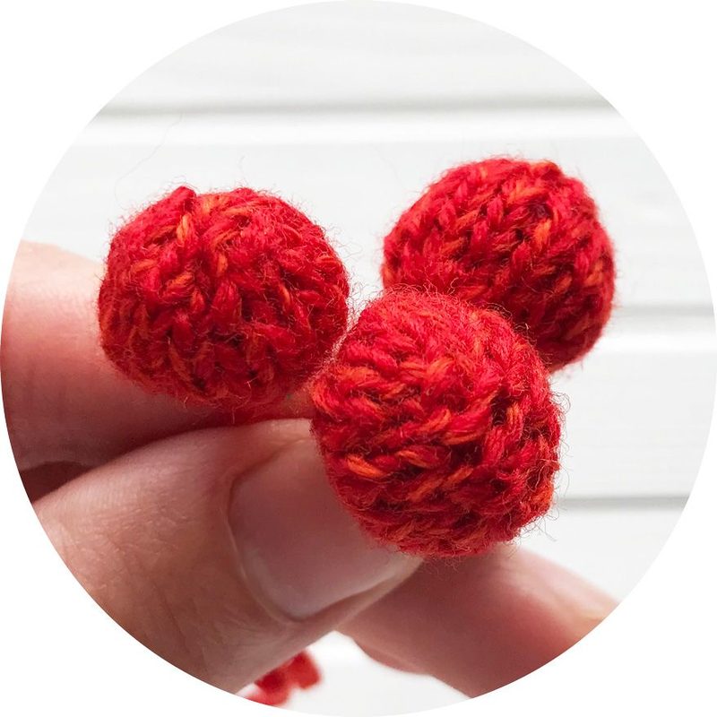 A close-up view of three small, knitted pom-poms held in a hand. The pom-poms are made of vibrant red yarn, with subtle variations in color that create a textured look. The background is softly blurred, ensuring the focus remains on the pom-poms. The image is framed in a circular format, highlighting the details of the knitting.
