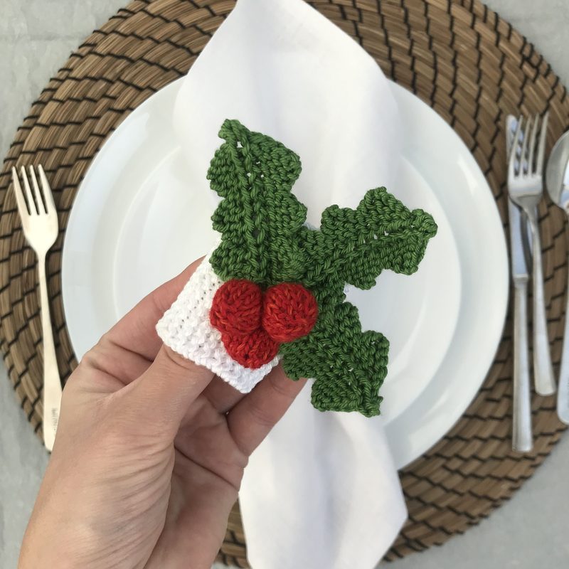 A person's hand holds a knitted napkin ring resembling holly, featuring dark green leaves and two red berries, with a white base. The napkin ring is set against a background of a round woven placemat and a white plate, which is partially visible. A folded white napkin is laid on the plate, with silver forks placed beside it. The overall color scheme includes green, red, and white, creating a festive appearance.