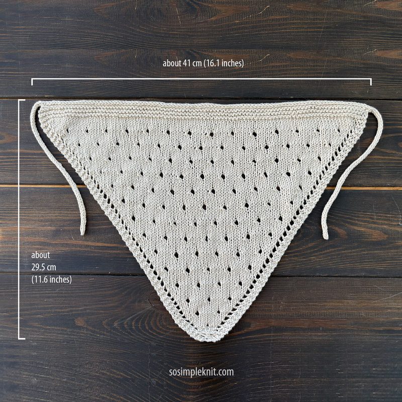A light beige knitted triangular scarf is displayed flat on a dark wooden surface. The scarf features a diamond-shaped lace pattern with evenly spaced holes, creating a delicate texture. It has two long ties at the pointed end for securing it around the neck. The dimensions are noted in inches along the edges, indicating the scarf measures about 41 cm (16.1 inches) in width and 29.5 cm (11.6 inches) in height. The overall look is minimal and elegant, emphasizing the simplicity of the knit design