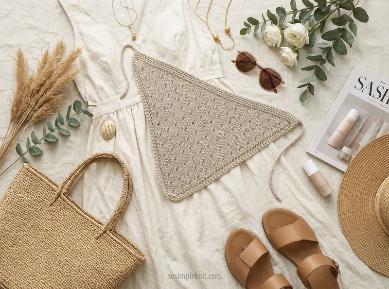 A knitted triangle scarf in a soft beige color is prominently displayed on a light, textured fabric background. Surrounding it are decorative elements, including a woven straw bag, brown sandals, a magazine titled "SASHA," sunglasses, and a small assortment of skincare products. Green eucalyptus leaves and white flowers are artfully arranged nearby, adding a natural touch to the composition. The overall scene conveys a casual and stylish aesthetic.