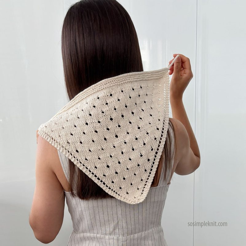 A person with long brown hair is shown from the back, wearing a cream-colored knitted triangle scarf with a dotted pattern. The scarf is draped around their shoulders, and the background appears to be a light-colored wall or curtain, creating a soft and neutral backdrop. The edges of the scarf have a textured finish, and the overall look is casual and feminine.