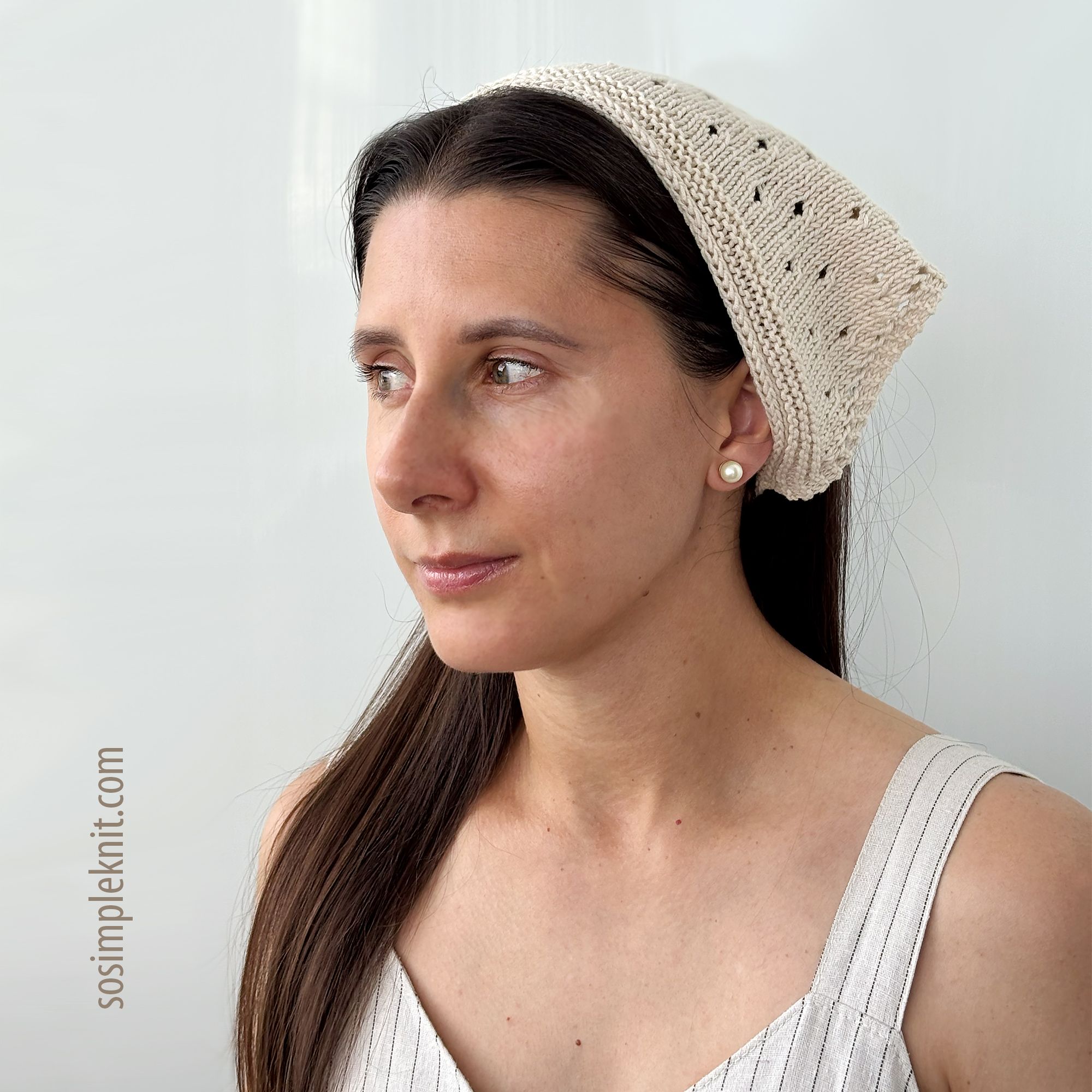 A woman with long brown hair wearing a cream-colored knitted bandana-style triangle scarf, featuring a lace pattern. The scarf drapes over her head and down the back, complementing her light-colored, sleeveless top. The background is a soft, neutral tone, enhancing the simplicity of the overall look. Her expression is calm and focused, with soft lighting illuminating her face.