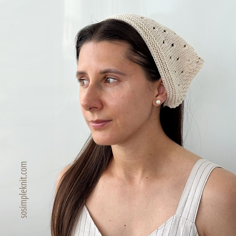A woman with long brown hair wearing a cream-colored knitted bandana-style triangle scarf, featuring a lace pattern. The scarf drapes over her head and down the back, complementing her light-colored, sleeveless top. The background is a soft, neutral tone, enhancing the simplicity of the overall look. Her expression is calm and focused, with soft lighting illuminating her face.