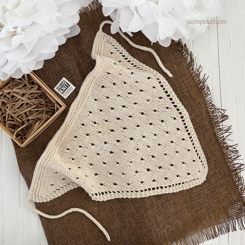 A knitted triangular bandana scarf in a creamy beige hue, featuring an openwork hole pattern throughout its body. The scarf is displayed on a textured brown woven fabric, complemented by a small square box containing straw-like material. The setting is minimalistic, with soft white flowers positioned in the background, creating a contrast with the earthy tones. The scarf's edges are neatly finished, emphasizing the craftsmanship of the knit design.