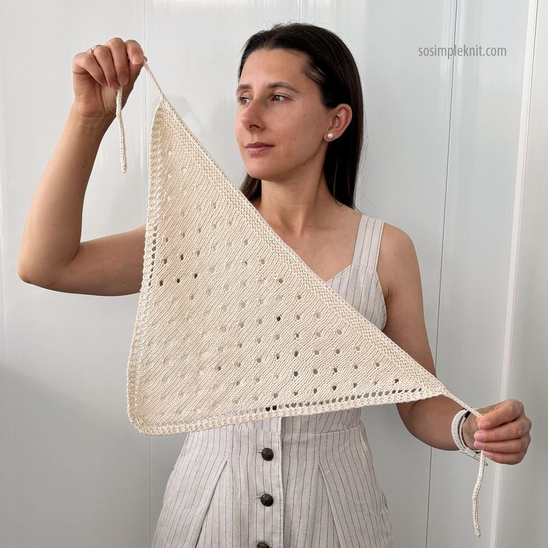 A woman stands against a plain white background, holding a triangular knitted bandana scarf made of light beige yarn. The scarf features an openwork pattern with small holes throughout, and she is showcasing it by holding one corner with her left hand while the other corner drapes down. She is wearing a light-colored, vertically striped dress and has her hair styled straight. Her expression is focused, drawing attention to the scarf.