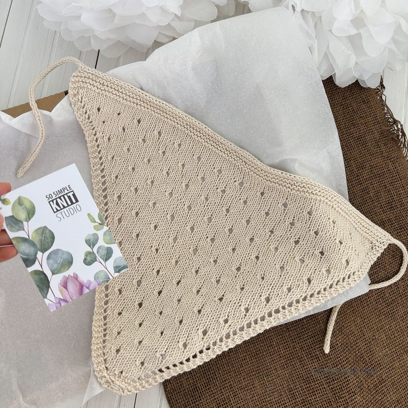 A light beige knitted bandana-style triangle scarf is displayed on a textured brown surface, with a piece of white tissue paper underneath. The scarf features a decorative openwork pattern with small holes and ties at the corners. A business card from "So Simple Knit Studio," showcasing muted green leaves and the studio name, is held beside the scarf, partially visible. The background includes soft white tissue paper and a neutral setting.
