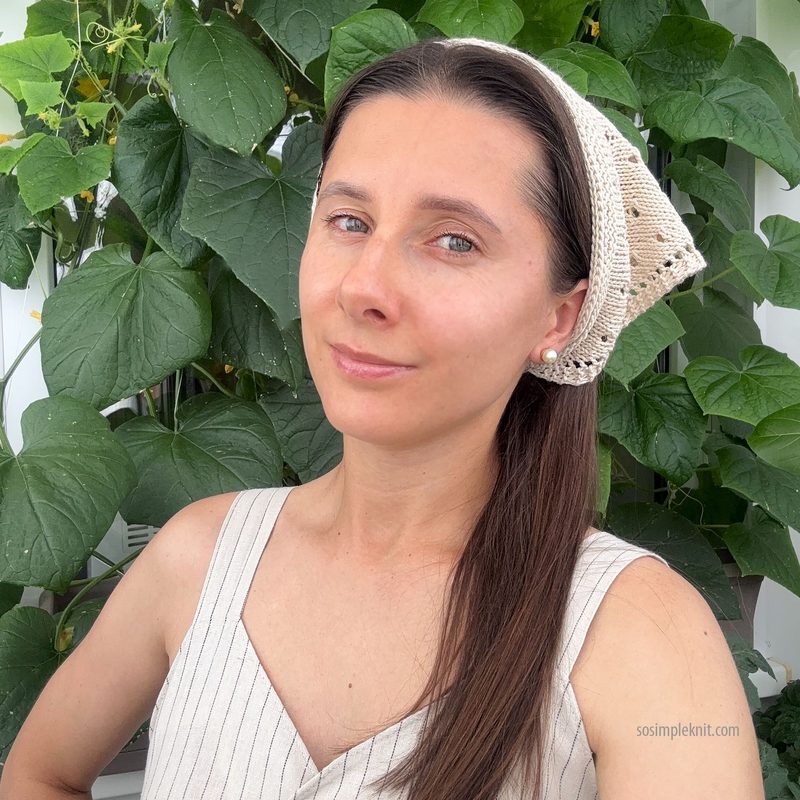 A woman with long brown hair is wearing a knitted triangle scarf as a bandana, featuring a textured pattern in light beige with dark spots along the edges. She is posing against a backdrop of green foliage, dressed in a sleeveless, striped top. The image is framed to show her upper body and face, with natural light illuminating her features.