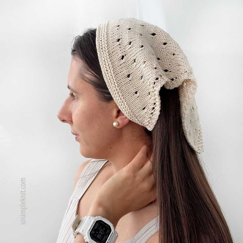 A woman is shown in profile wearing a knitted bandana-style triangle scarf in a light beige color. The scarf features an openwork pattern with small holes, allowing for a lightweight appearance. The woman's long dark hair is partially tucked into the scarf, and she is accessorized with a single pearl earring. The background is a soft, neutral color, emphasizing the scarf's texture and design. A watch is visible on her wrist.