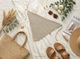 A knitted triangle scarf in a soft beige color is prominently displayed on a light, textured fabric background. Surrounding it are decorative elements, including a woven straw bag, brown sandals, a magazine titled "SASHA," sunglasses, and a small assortment of skincare products. Green eucalyptus leaves and white flowers are artfully arranged nearby, adding a natural touch to the composition. The overall scene conveys a casual and stylish aesthetic.