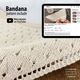The image shows a close-up of a knitted bandana pattern in a light beige color, with a triangular scarf shape highlighted in textured stitch details. In the background, a tablet displays a YouTube video tutorial related to the pattern, featuring a paused image of the knitting process. The overall composition focuses on the scarf and the instructional content, with a soft, neutral aesthetic.