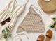 The image displays a knitted triangular bandana scarf in a light beige color with a textured pattern. Surrounding the scarf are a white linen dress, a pair of brown sandals, a woven basket bag, sunglasses, a gold necklace, a single white flower, and eucalyptus leaves. The arrangement is styled on a light, neutral background, creating a soft, airy aesthetic.