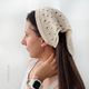 A woman is shown in profile wearing a knitted bandana-style triangle scarf in a light beige color. The scarf features an openwork pattern with small holes, allowing for a lightweight appearance. The woman's long dark hair is partially tucked into the scarf, and she is accessorized with a single pearl earring. The background is a soft, neutral color, emphasizing the scarf's texture and design. A watch is visible on her wrist.
