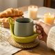 Lifestyle interior photo showing a cozy scene: a woman holds a cup in her hand while another cup rests on a knitted leaf-shaped coaster, demonstrating real-life use of the Leaf Coaster.