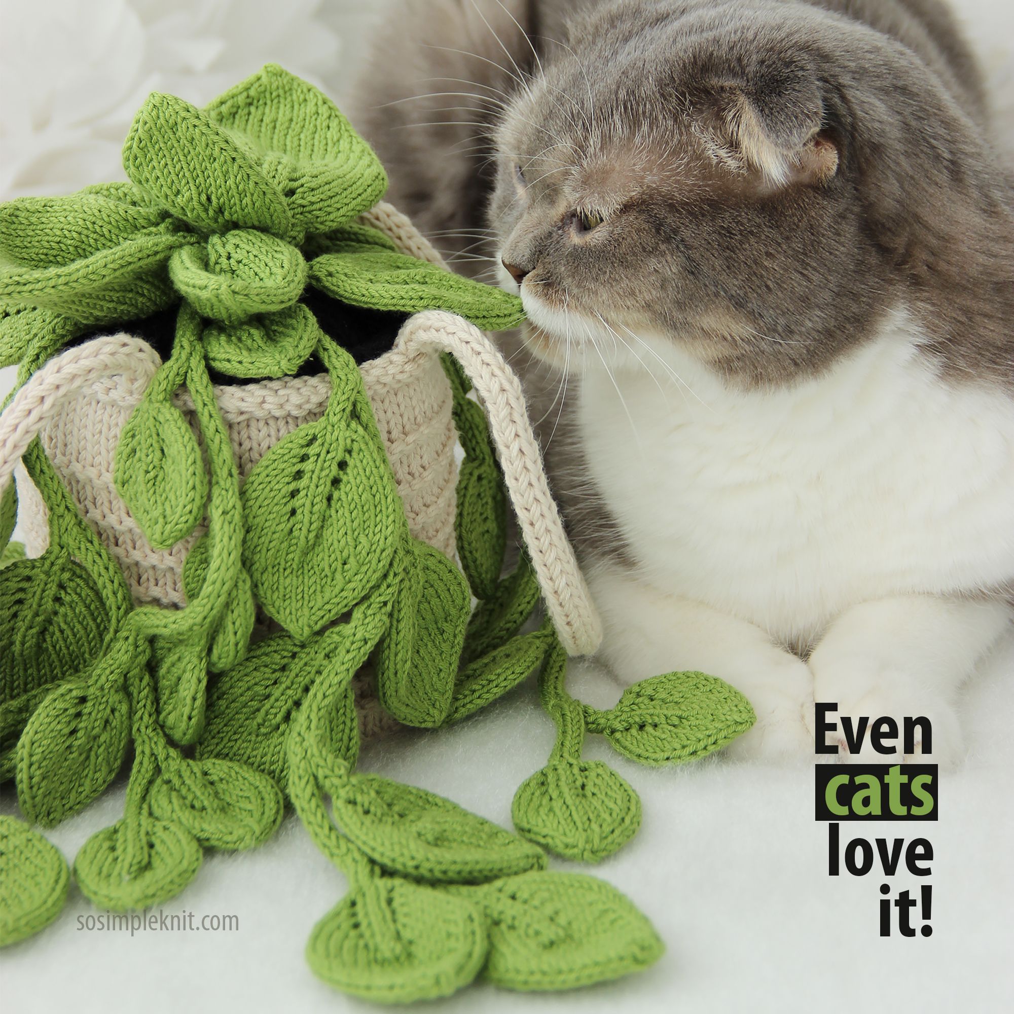 The image features a realistic knitted pothos plant with vibrant green leaves cascading out of a light beige pot. A fluffy gray and white cat is sitting beside the plant, looking curiously at it. The background is softly blurred, emphasizing the plant and the cat. In the lower right corner, there is bold text that reads “Even cats love it!” The overall style is cozy and inviting, showcasing the intricacy of the knitting and the charm of the cat.