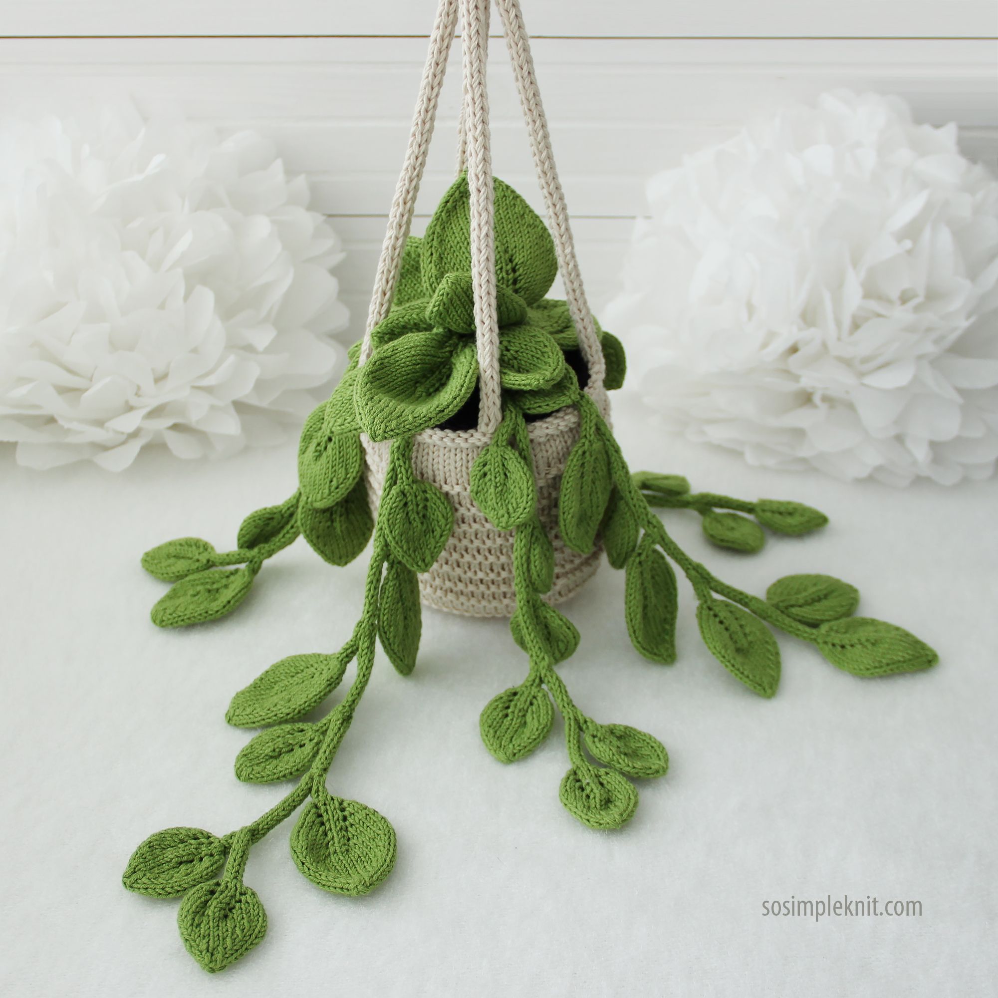 A knitted pothos plant is displayed in a beige, textured hanging basket, featuring long, trailing green leaves that cascade down. The leaves are varied in shape and size, showcasing a ribbed knitting pattern. The background consists of soft, white paper flowers, providing a contrasting touch to the greenery of the plant and the neutral tones of the basket. The image is well-lit, highlighting the details in both the plant and the basket.