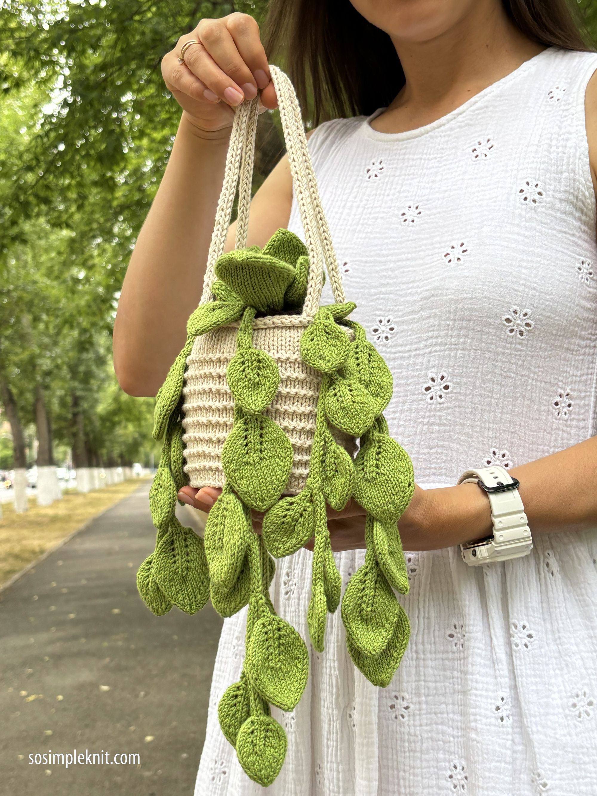 A person is holding a knitted pothos plant in a small cream-colored basket, which has a textured design. The plant features several vibrant green knitted leaves cascading down over the sides of the basket. The background includes blurred greenery and a tree-lined path, while the individual is wearing a white dress with a subtle pattern and a white watch.