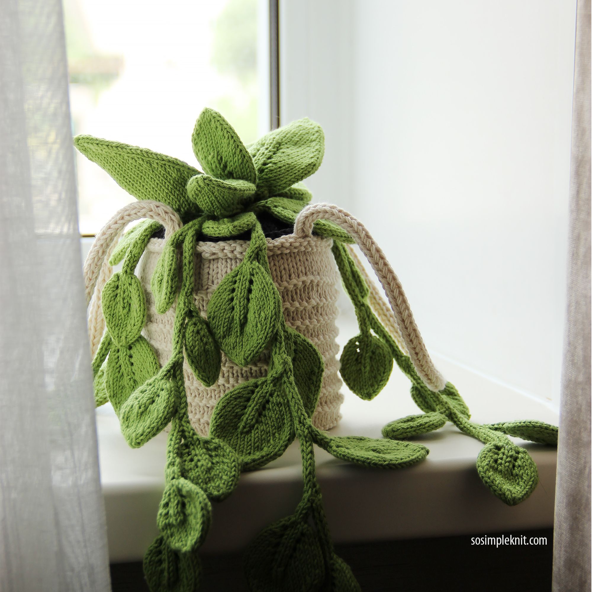 A knitted pothos plant sits in a textured cream-colored pot, with long green leaves cascading down. The leaves are vibrant and varied in shape, mimicking the appearance of a real pothos. The background features a softly lit window with sheer gray curtains that frame the scene, creating a calm and inviting atmosphere. The overall color palette includes shades of green and neutral tones, emphasizing the plant's lush appearance.
