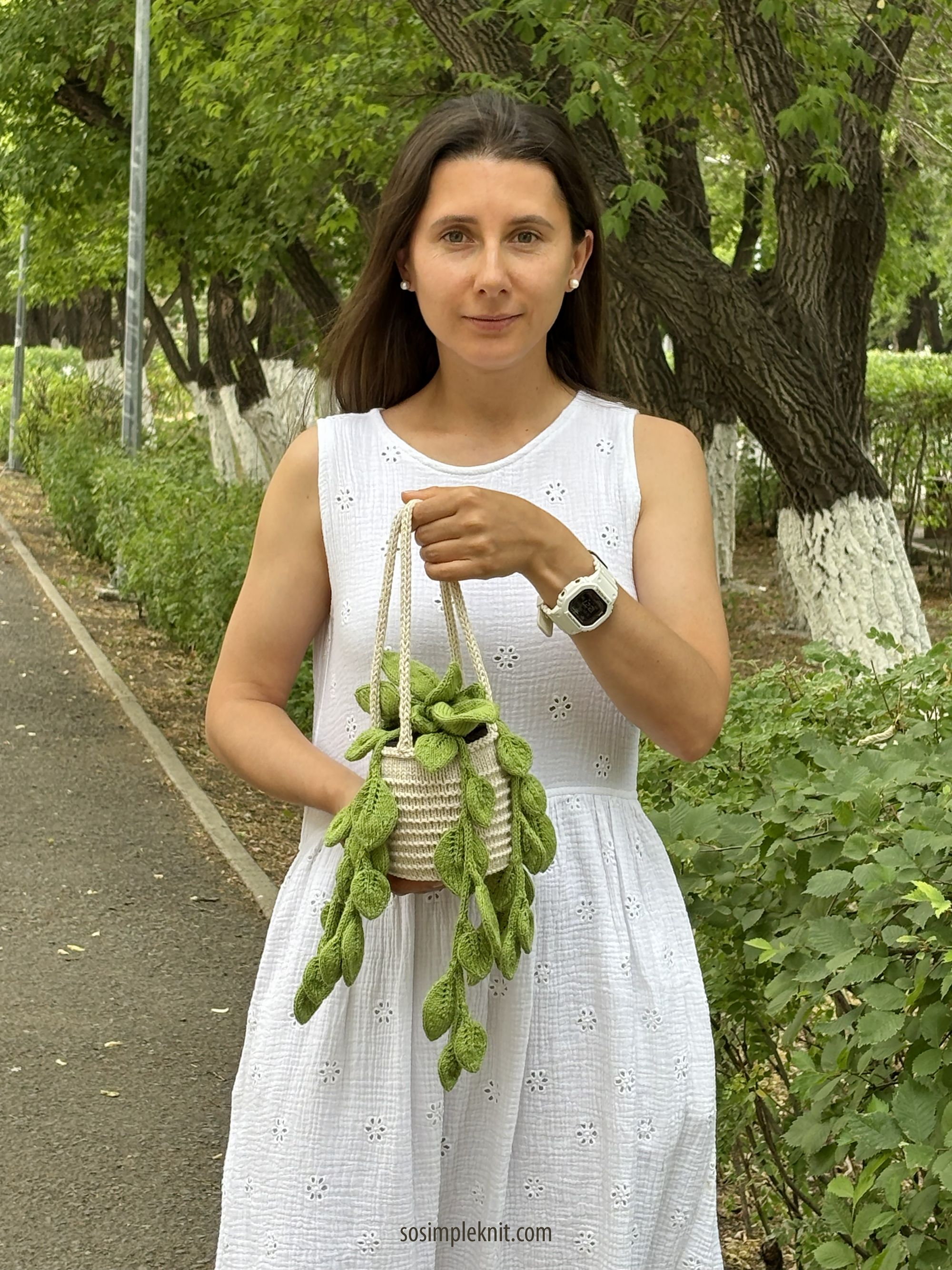 A woman stands on a pathway surrounded by greenery, holding a knitted pot adorned with green, leafy tendrils resembling a pothos plant. She wears a white, sleeveless dress with eyelet detailing and has long brown hair. The background features trees and a well-maintained path, creating a tranquil outdoor setting. The focus is on her and the knitted plant, with soft natural lighting enhancing the scene.