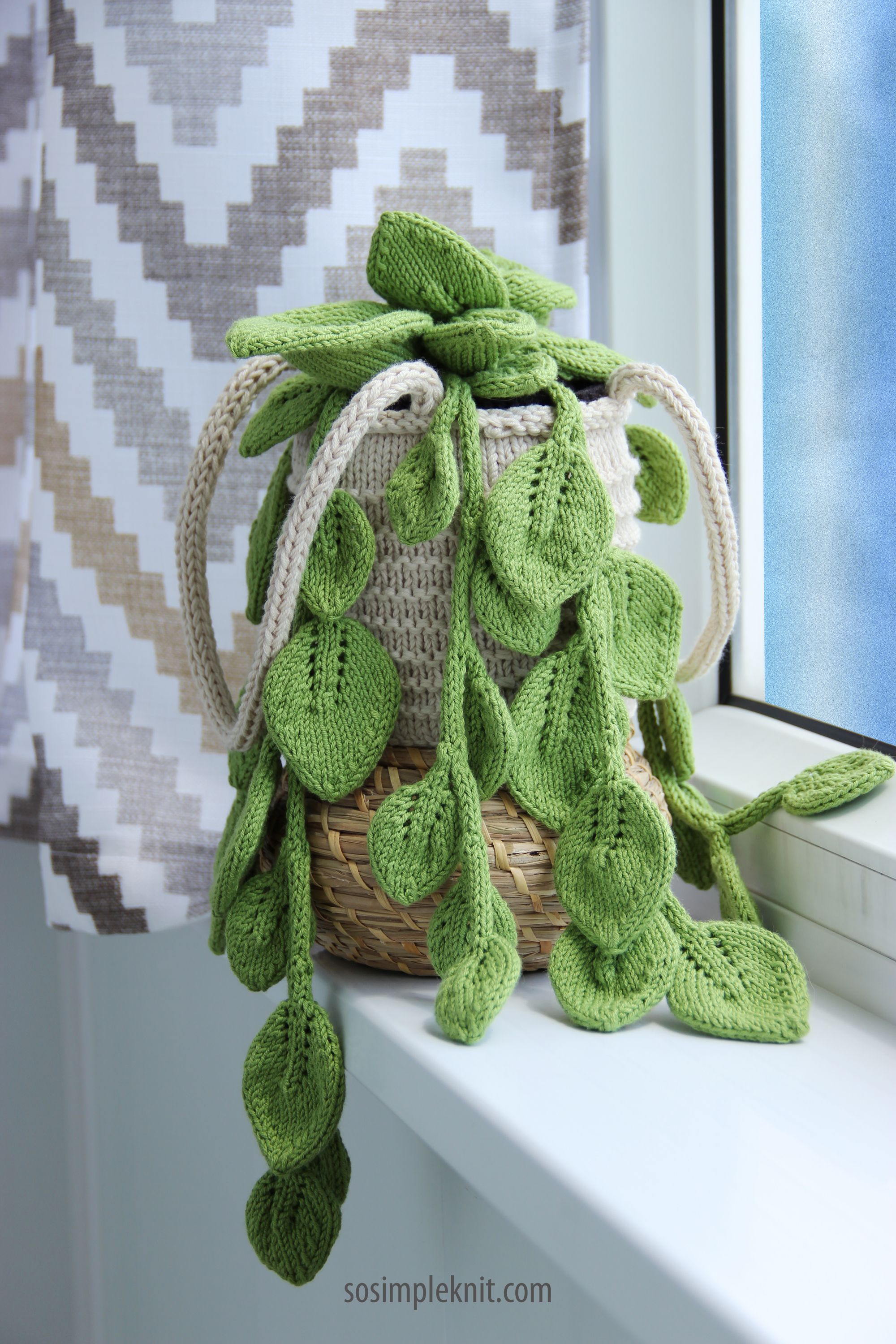 A knitted pothos hanging plant with large, textured green leaves cascades over the edge of a white and beige woven basket. The basket is placed on a window sill, with a white and gray chevron-patterned curtain in the background. Natural light illuminates the scene, highlighting the crafting details of the knitted leaves and the soft texture of the yarn.