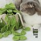 The image features a realistic knitted pothos plant with vibrant green leaves cascading out of a light beige pot. A fluffy gray and white cat is sitting beside the plant, looking curiously at it. The background is softly blurred, emphasizing the plant and the cat. In the lower right corner, there is bold text that reads “Even cats love it!” The overall style is cozy and inviting, showcasing the intricacy of the knitting and the charm of the cat.