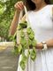 A person is holding a knitted pothos plant in a small cream-colored basket, which has a textured design. The plant features several vibrant green knitted leaves cascading down over the sides of the basket. The background includes blurred greenery and a tree-lined path, while the individual is wearing a white dress with a subtle pattern and a white watch.