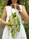 A person wearing a white dress holds a small, cream-colored knitted basket with a handle, filled with vibrant green knitted leaves resembling a pothos plant. The leaves cascade down from the basket, showing varying shades of green, and the background features a blurred outdoor setting with trees. The person's hand is positioned near the basket, highlighting the craftsmanship of the knitted elements.