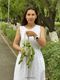 A woman stands on a pathway surrounded by greenery, holding a knitted pot adorned with green, leafy tendrils resembling a pothos plant. She wears a white, sleeveless dress with eyelet detailing and has long brown hair. The background features trees and a well-maintained path, creating a tranquil outdoor setting. The focus is on her and the knitted plant, with soft natural lighting enhancing the scene.
