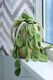 A knitted pothos hanging plant with large, textured green leaves cascades over the edge of a white and beige woven basket. The basket is placed on a window sill, with a white and gray chevron-patterned curtain in the background. Natural light illuminates the scene, highlighting the crafting details of the knitted leaves and the soft texture of the yarn.